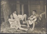 Two young men and a young woman sit with guitar relaxing together in the shade